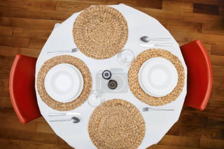 Top view of a round dining table set with white plates, silverware, woven placemats, and red chairs, ready for a meal.