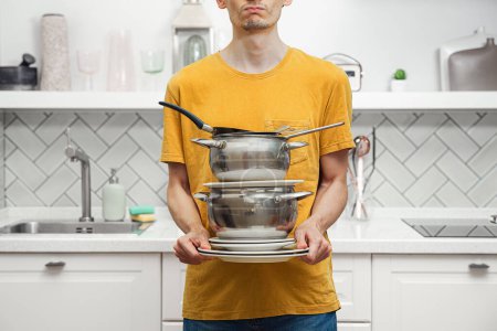 Foto de Equilibrio masculino con una pila de utensilios de cocina sucios y usados, platos, sartenes en el fondo de la cocina moderna, limpieza del hogar - Imagen libre de derechos