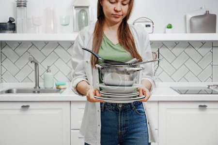 Müde Frau hält einen Stapel schmutziges Geschirr zum Waschen vor dem Hintergrund moderner Küche, routinemäßige Aufräumarbeiten nach den Mahlzeiten