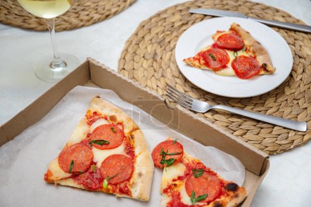 Photo for Casual dining setup featuring slices of pepperoni pizza on a plate and in a takeout box, paired with a glass of white wine. - Royalty Free Image