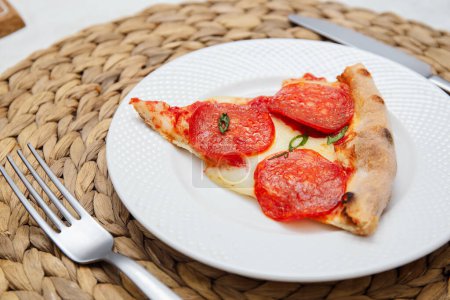 A single slice of pepperoni pizza with fresh basil on a white plate over a wicker placemat.