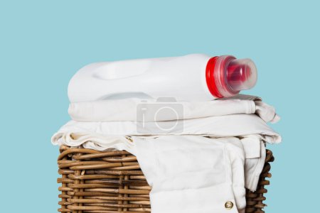 Eine Flasche Waschmittel neben einem Stapel frisch gewaschener weißer Kleidung in einem Weidenkorb vor blauem Hintergrund, frisch gereinigte und gefaltete Kleidung