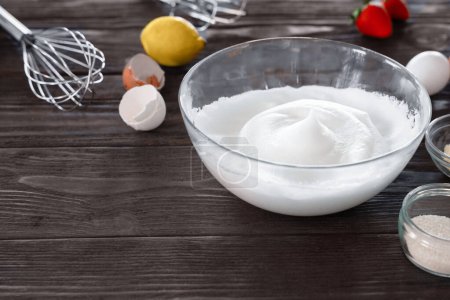 Bowl of whipped egg whites ready for baking, with a whisk, fresh lemon, and eggshells on a wooden countertop.