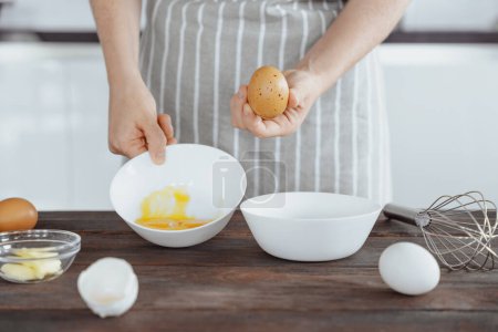 woman in apron separate egg whites from yolks in bowl for whisking or baking, home kitchen background, ingredient preparation
