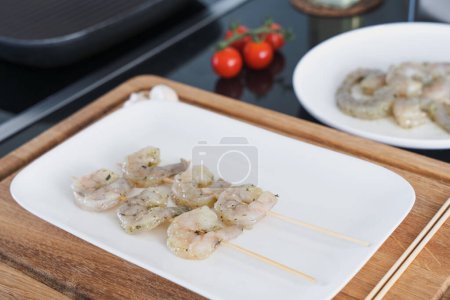 pinchos de camarón crudo marinado en un plato blanco, listo para asar en placa blanca y tabla de cortar de madera,
