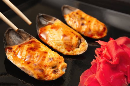 Three baked mussels topped with spicy mayonnaise, tobiko and teriyaki, served alongside pickled ginger on a sleek black plate with chopsticks.