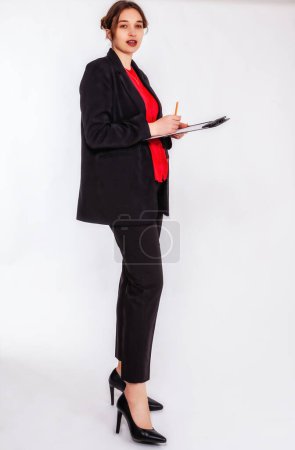 Una joven rubia con moño lleva un traje de pantalón negro y una blusa roja. Ella sostiene un tablero de escritura y un lápiz en su mano y mira atentamente a la cámara. El fondo es gris.