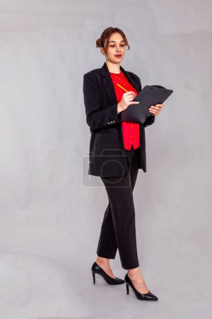 A young, blonde woman with a bun is wearing a black trouser suit and a red blouse. She holds a writing board and a pencil in her hand and looks intently into the camera. The background is gray.