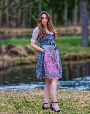 A young woman with long blonde hair stands in a traditional dirndl dress in a meadow. An idyllic stream flows behind her and a dense forest stretches out behind it. The woman smiles at the camera and enjoys the sweet spring air.