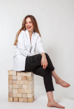 Una joven rubia de pelo largo se ríe en varios bloques de madera. Lleva una chaqueta blanca y pantalones negros. Sus pies están descalzos. La foto fue tomada en el estudio.
