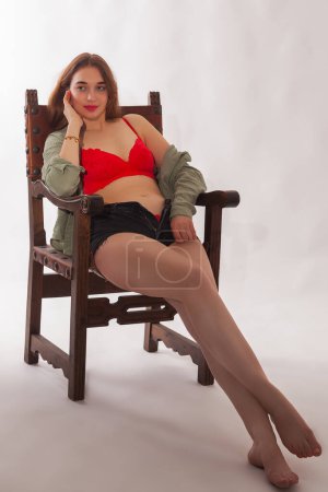 Una joven rubia de pelo largo está sentada en una silla española en un estudio. Ella está usando un conjunto de lencería roja que consiste en pantalones calientes y una parte superior de manga larga con un escote fuera del hombro. Ella se ve segura y sensual.