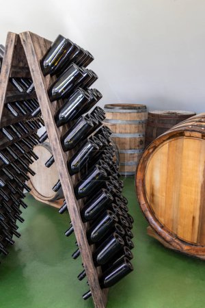 Photo for Wooden rack with empty wine bottles without labels, wooden wine barrels in the background - Royalty Free Image