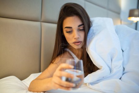 Photo for Young woman drinking glass of water in bed at night. Woman drinking a glass of water before going to sleep, she is lying in bed - Royalty Free Image