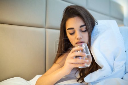 Photo for Young woman drinking glass of water in bed at night. Woman drinking a glass of water before going to sleep, she is lying in bed - Royalty Free Image