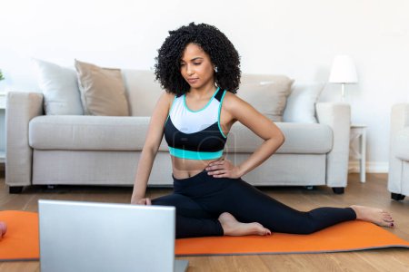 Foto de Una mujer africana adulta hace ejercicios de yoga y entrenamiento de fuerza en una estera en su sala de estar. Ella sigue un video del curso de ejercicio en línea en su computadora portátil. - Imagen libre de derechos