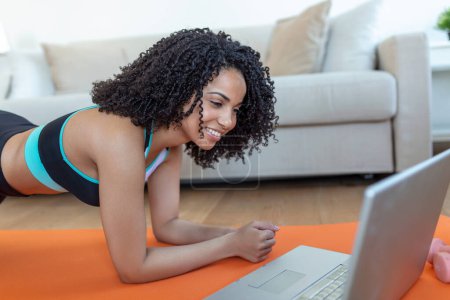 Foto de Una mujer africana adulta hace ejercicios de yoga y entrenamiento de fuerza en una estera en su sala de estar. Ella sigue un video del curso de ejercicio en línea en su computadora portátil. - Imagen libre de derechos