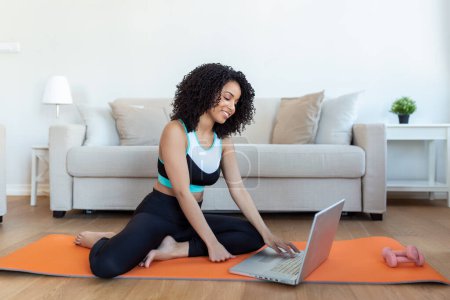 Téléchargez les photos : Une femme africaine adulte fait du yoga et des exercices de musculation sur un tapis dans son salon. Elle suit un cours d'exercice en ligne vidéo sur son ordinateur portable. - en image libre de droit