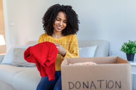 Mujer joven africana feliz sentarse en el sofá pegado ropa en la caja de donaciones en casa, cuidado biracial voluntario poner ropa en el paquete de cartón, donar a las personas necesitadas, reutilizar, reciclar el concepto