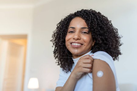 Foto de Portrait of a female smiling after getting a vaccine. Woman holding down her shirt sleeve and showing her arm with bandage after receiving vaccination. - Imagen libre de derechos