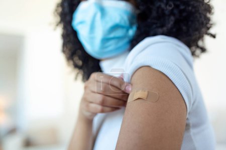 Photo for African woman holding up her shirt sleeve and showing her arm with bandage after receiving vaccination. covid 19 immunization - Royalty Free Image