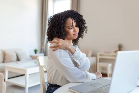 Téléchargez les photos : Portrait de jeune femme stressée assise au bureau à domicile devant un ordinateur portable, touchant le dos avec une expression douloureuse, souffrant de maux de dos après avoir travaillé sur PC - en image libre de droit