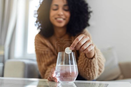 Téléchargez les photos : Closeup of a young woman dropping an effervescent antacid in a glass of water. young woman hardly put a soluble pill with a medicine for pain or a hangover in a glass of water - en image libre de droit