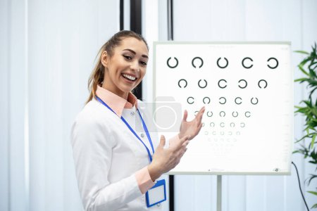 Photo for Professional female optician pointing at eye chart, timely diagnosis of vision. Portrait of optician asking patient for an eye exam test with an eye chart monitor at his clinic - Royalty Free Image