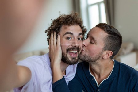 Téléchargez les photos : Beau gay assis ensemble à la maison prenant selfie avec téléphone mobile. gay l'homme avec son partenaire assis en prenant un auto portrait à l'aide de téléphone intelligent. - en image libre de droit