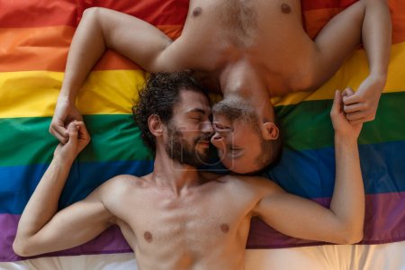 Hombre gay joven cariñoso besando a su amante en la cama. Dos jóvenes amantes varones sentados juntos en la bandera del orgullo. Romántico joven gay pareja vinculación cariñosamente en el interior.