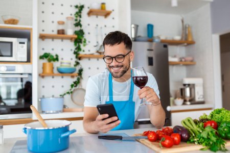 Photo for Head shot smiling young man using smartphone, chatting in social network while preparing food for vegetarian dinner at home. Happy millennial guy in eyeglasses web surfing recipe for meal in kitchen. - Royalty Free Image