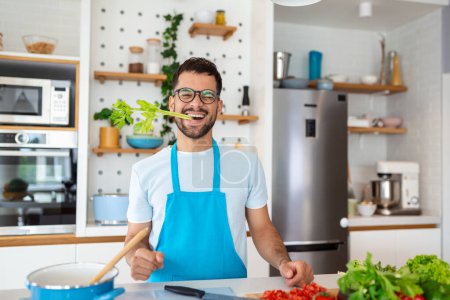 Foto de Feliz hombre guapo cocinando en la cocina en casa con apio en la boca posando para la foto, concepto de vida saludable - Imagen libre de derechos