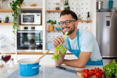 Photo for Handsome smiling young man leaning on kitchen counter with vegetables and looking away. Portrait of happy casual guy in apron leaning on steel counter in the kitchen with ingredients on it. - Royalty Free Image