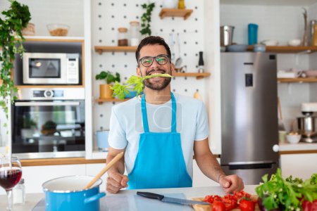Téléchargez les photos : Joyeux bel homme cuisinant dans la cuisine à la maison avec du céleri dans la bouche posant pour la photo, concept de vie saine - en image libre de droit