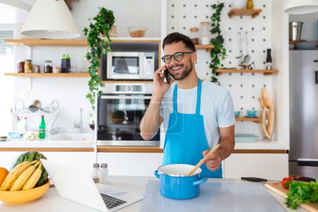 Foto de Atractivo joven está cocinando en la cocina con el ordenador portátil en la mesa mientras habla en el teléfono inteligente
. - Imagen libre de derechos