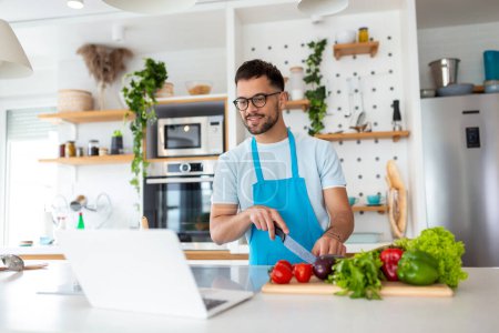 Photo for Happy young man cooking healthy dinner at home. He is following a video tutorial on the laptop. - Royalty Free Image