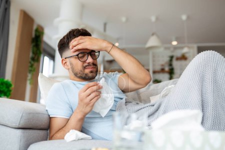Photo for Man feeling sick lying in the bed and looking the thermometer. Sick man lying on sofa checking his temperature under a blanket at home in the living room - Royalty Free Image