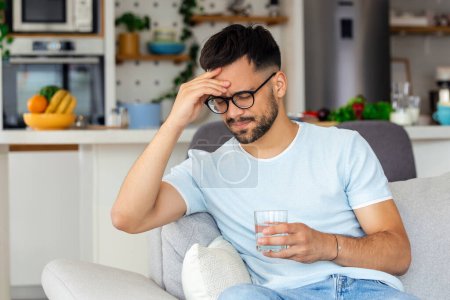 Photo for Young man suffering from strong headache or migraine sitting with glass of water on the sofa, millennial guy feeling intoxication and pain touching aching head, morning after hangover concept - Royalty Free Image