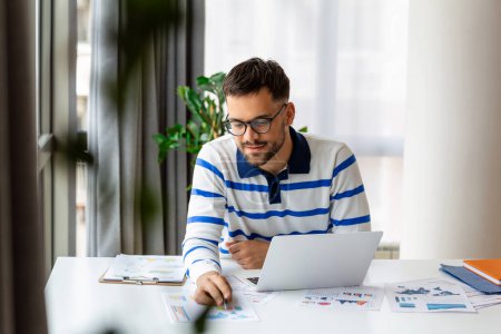 Téléchargez les photos : Homme souriant dans des lunettes assis au bureau en regardant les données tout en travaillant sur son ordinateur portable - en image libre de droit