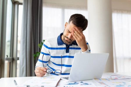 Photo for Young businessman touching his forehead while working on laptop at home. Portrait of young man having severe headache while sitting at home table and working on computer - Royalty Free Image