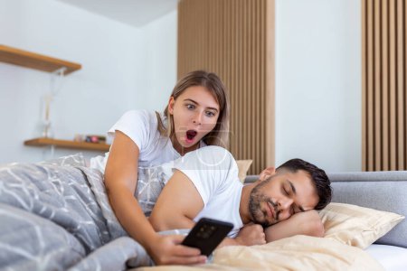 Téléchargez les photos : Petite amie jalouse espionnant le téléphone de son partenaire pendant qu'il dort dans un lit à la maison. Choqué femme jalouse espionnage le téléphone de son mari pendant qu'il dort - en image libre de droit