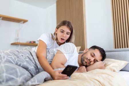 Photo for Jealous wife spying the phone of her partner while he is sleeping in a bed at home. Shocked jealous wife spying the phone of her husband while man sleeping in bed at home - Royalty Free Image
