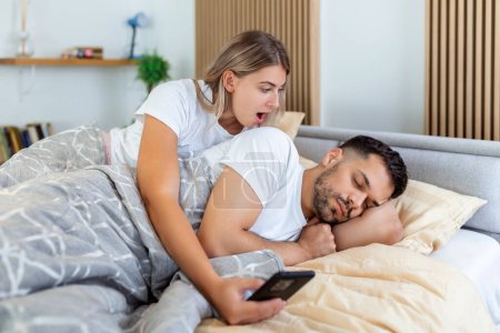 Photo for Woman is jealous and suspicious and spies in her partner's smartphone whiles he's sleeping in bedroom. The wife is spying on her husband's phone while he sleeps. The concept of distrust, jealousy - Royalty Free Image
