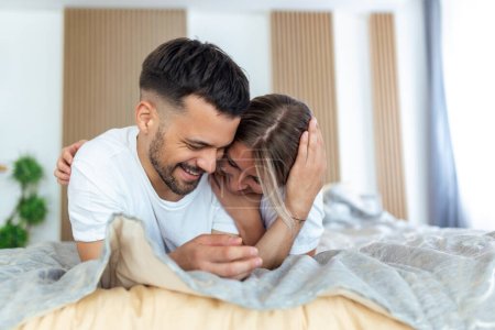 Téléchargez les photos : Couple souriant va embrasser sous les draps de couchage le matin. Dans l'amour jeune couple heureux couché dans le lit le matin - en image libre de droit