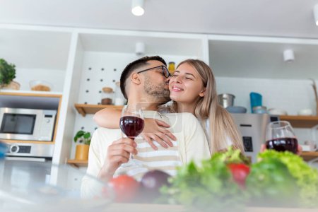 Photo for Affectionate young man kissing his wife while cooking together. Beautiful young couple is talking and smiling while cooking healthy food in kitchen at home.Man is kissing his girlfriend in cheek - Royalty Free Image
