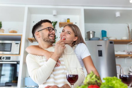 Téléchargez les photos : Beau jeune couple se regarde et se nourrir avec des sourires tout en cuisinant dans la cuisine à la maison. Amour joyeux jeune couple embrasser et cuisiner ensemble, s'amuser dans la cuisine - en image libre de droit