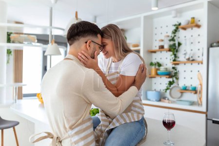 Photo for Affectionate young man kissing his wife while she's making breakfast. Beautiful young couple is talking and smiling while cooking healthy food in kitchen at home.Man is kissing his girlfriend in cheek - Royalty Free Image