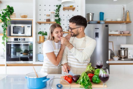 Photo for Beautiful young couple is looking at each other and feeding each other with smiles while cooking in kitchen at home. Loving joyful young couple embracing and cooking together, having fun in the kitchen - Royalty Free Image
