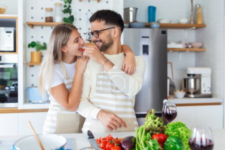 Téléchargez les photos : Beau jeune couple se regarde et se nourrir avec des sourires tout en cuisinant dans la cuisine à la maison. Amour joyeux jeune couple embrasser et cuisiner ensemble, s'amuser dans la cuisine - en image libre de droit