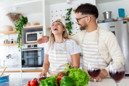 Photo for Beautiful young couple is feeding each other and smiling while cooking in kitchen at home. Happy sporty couple is preparing healthy food on light kitchen. Healthy food concept. - Royalty Free Image