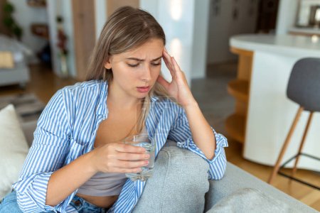 Foto de Mujer joven deprimida con dolor de cabeza mientras bebe agua en el sofá en casa. - Imagen libre de derechos